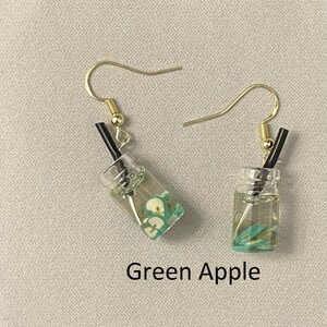 May include: A pair of dangle earrings featuring miniature clear glass beverage cups with green apple slices and black straws. The earrings have gold-colored fish hook ear wires. The text "Green Apple" is below the earrings.