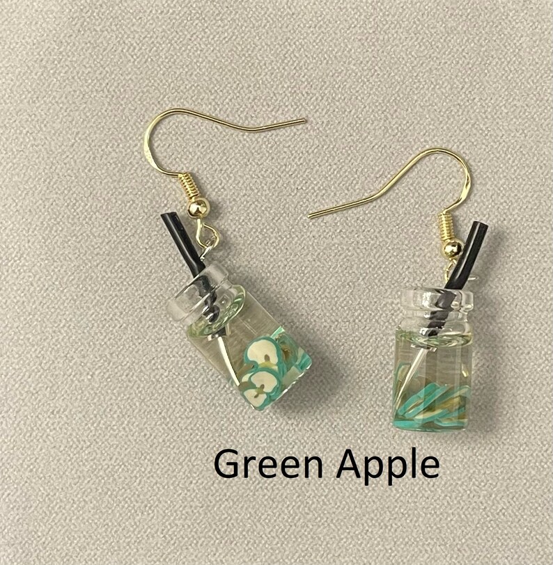 May include: A pair of dangle earrings featuring miniature clear glass beverage cups with green apple slices and black straws. The earrings have gold-colored fish hook ear wires. The text "Green Apple" is below the earrings.