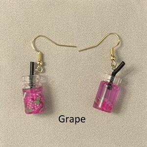 May include: A pair of gold-colored hook earrings. Each earring features a miniature clear plastic cup filled with purple liquid and small strawberry-shaped pieces. A black straw extends from the top of each cup. The word "Grape" is printed below the earrings.