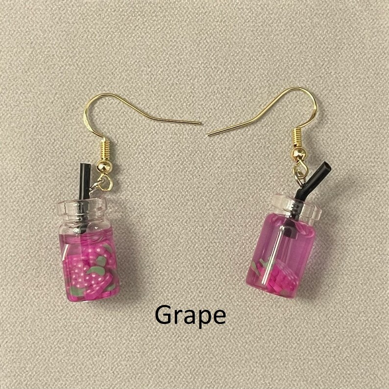 May include: A pair of gold-colored hook earrings. Each earring features a miniature clear plastic cup filled with purple liquid and small strawberry-shaped pieces. A black straw extends from the top of each cup. The word "Grape" is printed below the earrings.