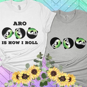 Puede incluir: Dos camisetas, una blanca y otra gris, con el texto "ARO IS HOW I ROLL". Cada camiseta presenta tres caras de panda con los colores de la bandera del orgullo arromántico. Arreglo floral de girasoles y vegetación en la parte inferior.