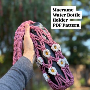 Peut inclure: Un porte-bouteille d'eau en macramé rose avec des accents de marguerites blanches. Le support est conçu pour transporter une bouteille d'eau noire. Le texte "Macrame Water Bottle Holder PDF Pattern" est visible.