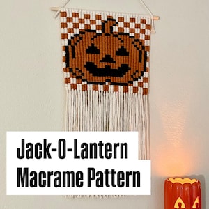 May include: A macrame wall hanging featuring a pixelated orange and black jack-o-lantern design set against a white and orange checkered background, suspended from a wooden dowel with long white fringe and the text Jack-O-Lantern Macrame Pattern.