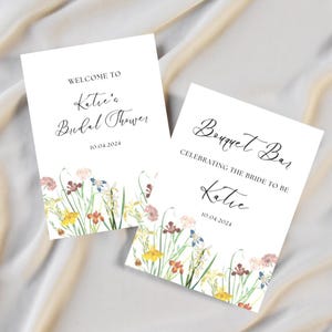 May include: Two white signs with black text and floral borders. The first sign says "Welcome to Katie's Bridal Shower 10.04.2024". The second sign says "Bouquet Bar Celebrating the Bride to be Katie 10.04.2024".