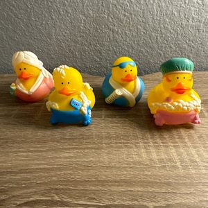 May include: Four yellow rubber duck toys with different accessories. One duck wears a pink robe and a white head wrap. Another has a blue tag that says "SOAP". A third duck wears blue swim goggles and a blue and white towel. The last duck is in a pink bathtub with a green cap.