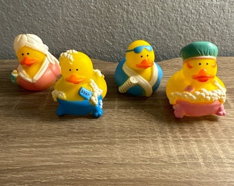 May include: Four yellow rubber duck toys with different accessories. One duck wears a pink robe and a white head wrap. Another has a blue tag that says "SOAP". A third duck wears blue swim goggles and a blue and white towel. The last duck is in a pink bathtub with a green cap.