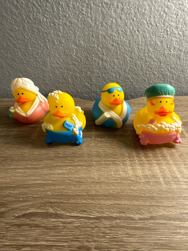 May include: Four yellow rubber duck toys with different accessories. One duck wears a pink robe and a white head wrap. Another has a blue tag that says "SOAP". A third duck wears blue swim goggles and a blue and white towel. The last duck is in a pink bathtub with a green cap.