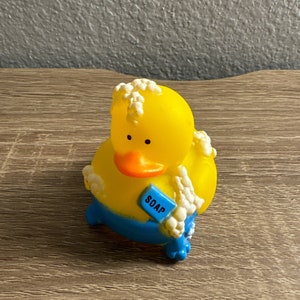 May include: A yellow rubber duck toy with a blue bottom and orange beak. The duck has white suds around its head and body and is holding a blue rectangle with the word "SOAP" on it. The toy is on a wooden surface.