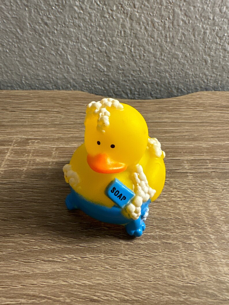 May include: A yellow rubber duck toy with a blue bottom and orange beak. The duck has white suds around its head and body and is holding a blue rectangle with the word "SOAP" on it. The toy is on a wooden surface.