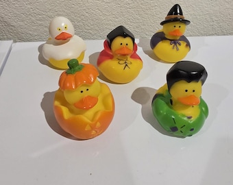 May include: A collection of five yellow rubber ducks dressed in Halloween costumes. One is a ghost, one a vampire, one a witch, one inside a jack-o'-lantern, and one wearing a green monster-themed outfit. Each duck has a bright orange beak.
