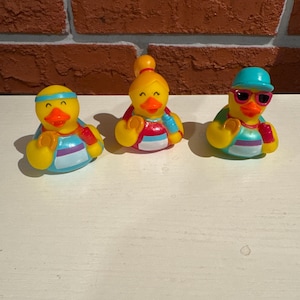 May include: Three yellow rubber duck toys with different accessories. One duck wears a blue headband, another has a topknot, and the third sports sunglasses and a blue cap. Each duck has a striped shirt in shades of blue, red, and white.
