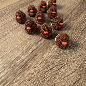May include: A group of brown beaded chick-shaped ornaments with orange beaks and white detailing. The ornaments are displayed on a wooden surface. The beaded texture and design suggest a craft or decorative item.