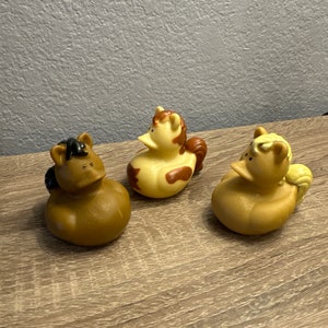 May include: Three rubber duck toys shaped like horses. The ducks are brown, tan, and yellow with black, brown, and yellow accents.