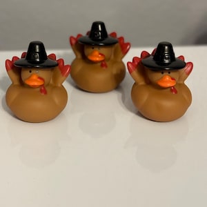 May include: Three brown rubber turkey toys, each wearing a black pilgrim hat. The toys have orange beaks and red tail feathers. They are arranged on a white surface. A fun Thanksgiving decoration.