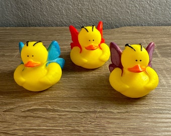 May include: Three yellow rubber duck toys with butterfly wings. The ducks have black eyes and a black stripe across their heads. The wings are different colors: red, blue, and purple.