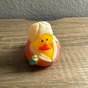 May include: A rubber ducky wearing a turban and a pink and orange robe, with a small teal and white accessory. The duck has a yellow body, orange beak, and black eyes. The duck is a novelty bath toy.
