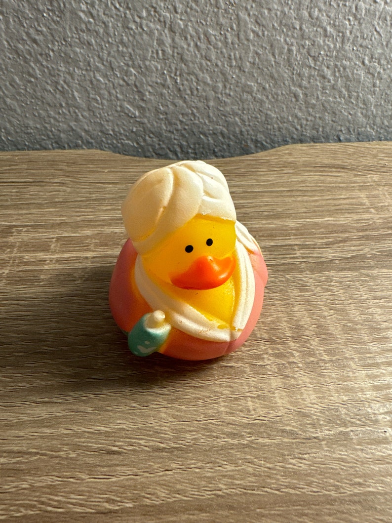 May include: A rubber ducky wearing a turban and a pink and orange robe, with a small teal and white accessory. The duck has a yellow body, orange beak, and black eyes. The duck is a novelty bath toy.