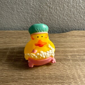 May include: A yellow rubber duck wearing a teal shower cap and a pink bathtub. The duck has an orange beak and is surrounded by white bubbles. The duck is a fun bath toy.