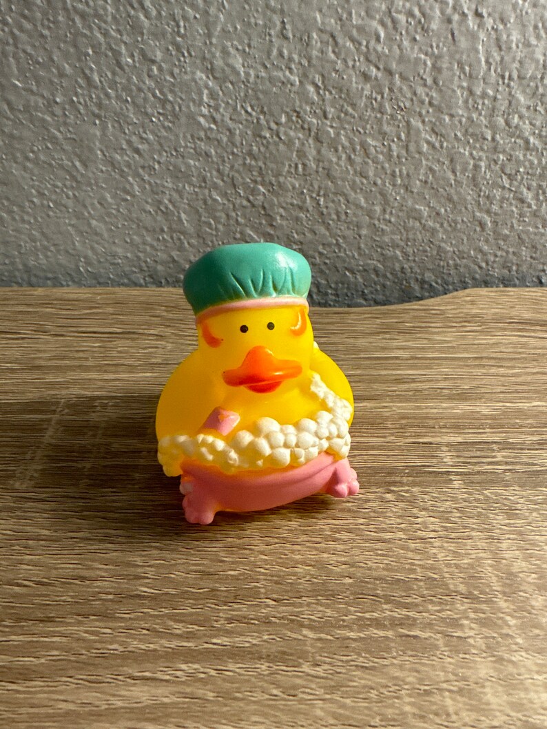 May include: A yellow rubber duck wearing a teal shower cap and a pink bathtub. The duck has an orange beak and is surrounded by white bubbles. The duck is a fun bath toy.
