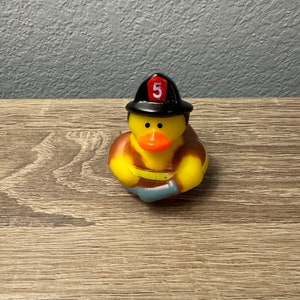 May include: A yellow rubber duck wearing a black firefighter hat with a red number 5 on it. The duck is holding a blue hose in its hands.
