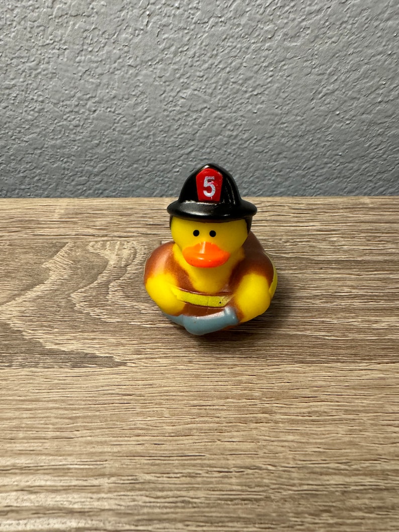 May include: A yellow rubber duck wearing a black firefighter hat with a red number 5 on it. The duck is holding a blue hose in its hands.
