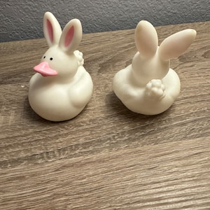 May include: Two white rubber duckies shaped like bunnies with pink ears. The duckies are sitting on a wooden surface.