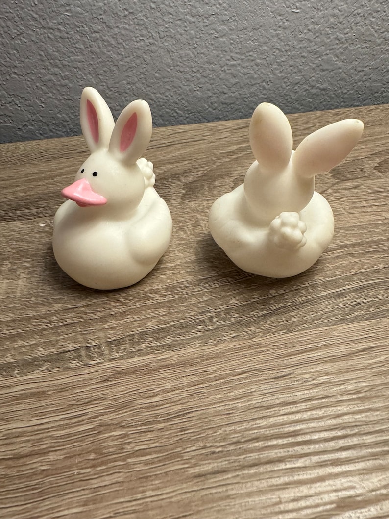 May include: Two white rubber duckies shaped like bunnies with pink ears. The duckies are sitting on a wooden surface.