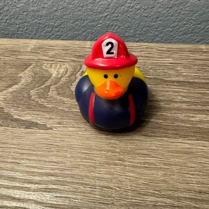 May include: Yellow rubber duck wearing a red firefighter hat with the number 2 on it. The duck is wearing a blue and red firefighter uniform.