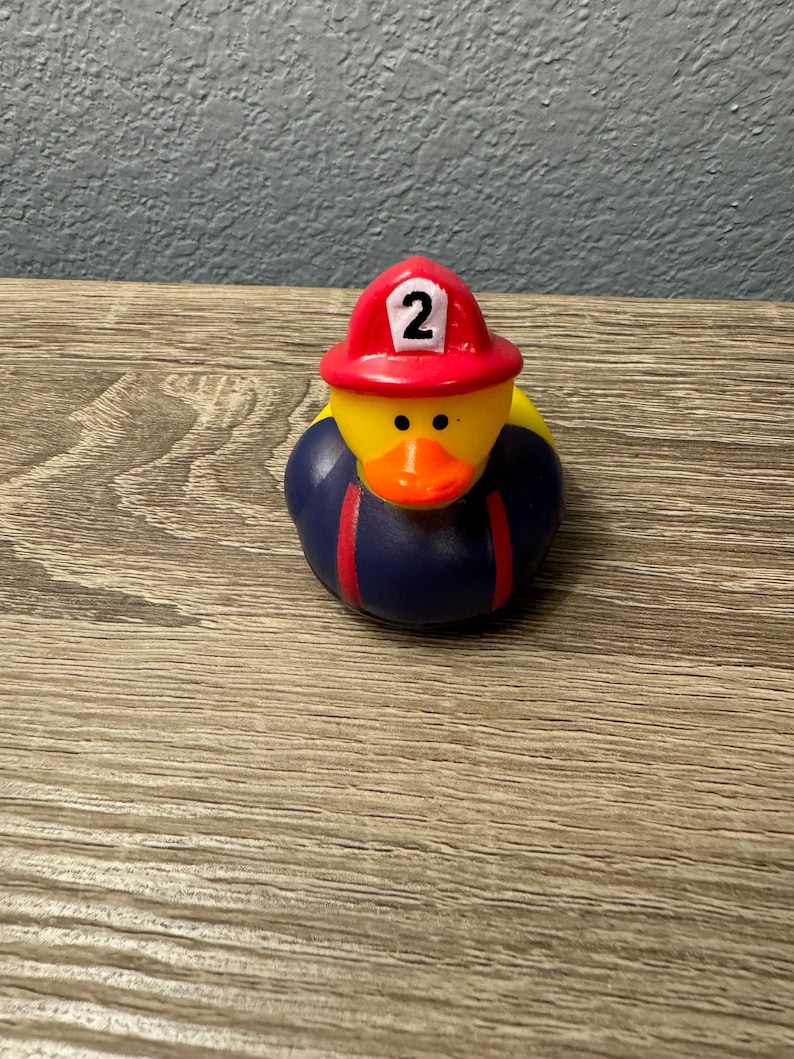 May include: Yellow rubber duck wearing a red firefighter hat with the number 2 on it. The duck is wearing a blue and red firefighter uniform.