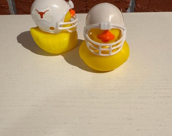 May include: Two yellow rubber duckies wearing white football helmets. One helmet has a red Longhorn logo. The ducks have orange beaks and are on a white surface. The background is a brick wall.