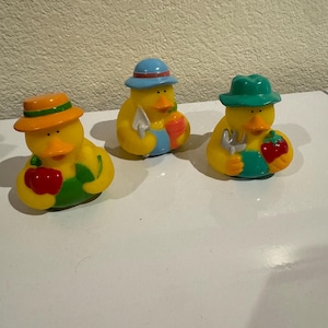 May include: Three yellow rubber ducks dressed as gardeners. The left duck wears an orange hat and green overalls holding a red apple. The middle duck wears a blue hat and blue overalls holding a trowel. The right duck wears a green hat and green overalls holding a silver garden fork and a red strawberry.