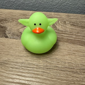 May include: A green rubber duck with large ears and orange beak. The duck is sitting on a wooden surface.