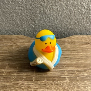 May include: A yellow rubber duck wearing blue swim goggles and a blue and white towel. The duck has an orange beak and is a fun, whimsical bath toy.
