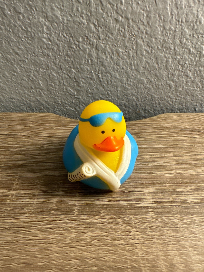 May include: A yellow rubber duck wearing blue swim goggles and a blue and white towel. The duck has an orange beak and is a fun, whimsical bath toy.