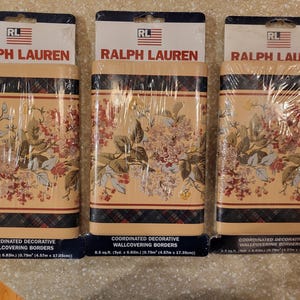 May include: Three rolls of Ralph Lauren coordinated decorative wallcovering borders. The wallpaper features a floral pattern in shades of pink, white, and green on a beige background. The borders are wrapped in clear plastic.
