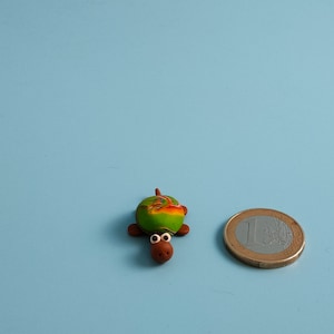 Op de afbeelding: Een kleine, handgemaakte schildpadfiguur met een groen en oranje schild, bruine ledematen en een bruine kop met wit omrande ogen. Naast de schildpad ligt een munt van één euro. De schildpad is ongeveer 2,5 cm lang.