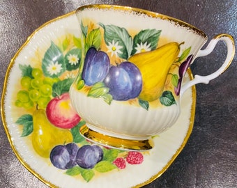 Op de afbeelding: Een wit theekopje en schotel met een gouden rand. Het kopje en de schotel zijn versierd met een kleurrijk fruitpatroon, waaronder peren, pruimen, druiven en frambozen.