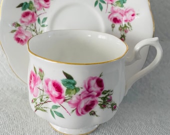 May include: A white teacup and saucer with a pink rose floral pattern. The cup and saucer have a gold trim around the edge.