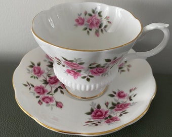 May include: A white teacup and saucer set decorated with pink rose floral patterns. The edges of the cup and saucer are scalloped and trimmed with gold. The teacup has a curved handle.