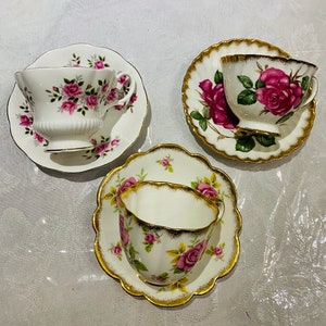以下が含まれることがあります： ピンクのバラの花柄と金色の縁取りが施された白いティーカップとソーサーが3つ。カップとソーサーは波打った縁をしています。