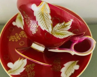 May include: A deep red porcelain teacup and saucer set featuring white leaf motifs with gold trim and delicate gold floral accents. The cup has a pedestal base and a curved handle, while the saucer displays matching leaf and circle patterns.