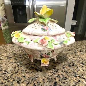 Puede incluir: Un plato de cerámica decorativo con tapa, con un adorno de rosa amarilla. El plato está adornado con detalles florales rosas, amarillos y verdes. La base tiene pequeños pies y está colocado sobre una encimera moteada.