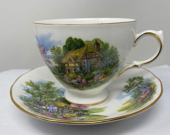 May include: A white teacup and saucer set with a gold rim. The teacup and saucer feature a colorful cottage scene with a thatched roof, green trees, and blue flowers.