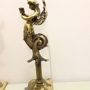 Op de afbeelding: Een messing kandelaar met een zeemeerminachtige figuur met vleugels, die een kleine beker vasthoudt. De figuur staat op een gegroefde kolom en een vierkante basis met sierlijke details. Het ontwerp is antiek en decoratief.