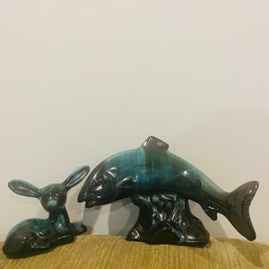 May include: Ceramic figurines of a rabbit and a fish. The rabbit is dark teal with long ears, lying down. The fish is teal and black, mounted on a black base. The figurines are on a green surface.