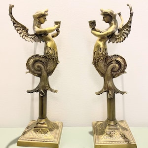 May include: A pair of antique brass candle holders. Each features a winged figure with a mermaid tail, holding a small cup. The figures are mounted on a square base with ornate detailing. The candle holders are approximately 30 cm tall.