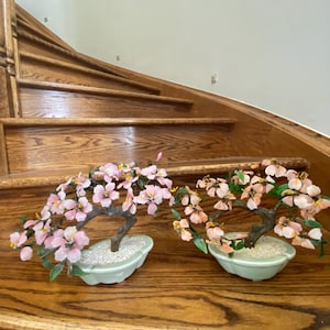 May include: Two glass bonsai trees with pink flowers in green ceramic pots. The trees are on a wooden staircase.