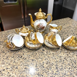 Könnte beinhalten: Ein Vintage-Teeservice mit goldenen Verzierungen und floralen Designs. Das Set umfasst eine Teekanne, eine Zuckerdose, einen Sahnekännchen und mehrere Teetassen mit Untertassen. Das Teeservice ist auf einer gesprenkelten Arbeitsplatte angeordnet.