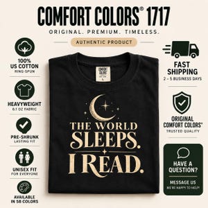 May include: A black short-sleeve t-shirt featuring a graphic print in a light beige color. The design includes a crescent moon, stars, and the text "THE WORLD SLEEPS. I READ." The shirt has a Comfort Colors brand label on the inside of the collar.