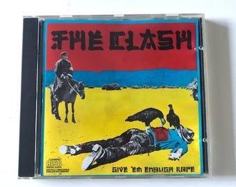 May include: CD case for The Clash's album "Give 'Em Enough Rope." The cover art features a red and blue sky over a yellow field, with a figure on horseback and a person lying on the ground with birds.